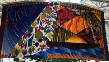 Colorful patchwork tapestry hanging from the ceiling, includes flying kites, leaves and flours, wheat, grain, rocks.